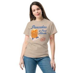 May include: A beige t-shirt with the words "Pancakes & Panic Attacks" in blue. The design shows a stack of pancakes with butter, blueberries, and a small "buy one get one" sticker.