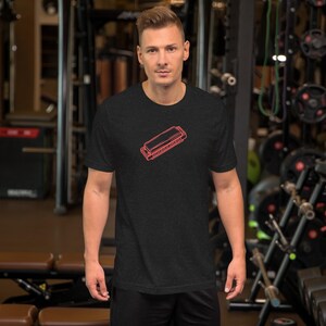 May include: Dark grey t-shirt with a red outline of a harmonica. The shirt has a crew neck and is made of a soft material. The design is a simple graphic of a musical instrument.