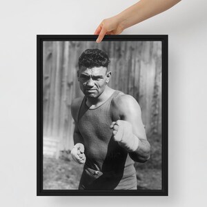 May include: Black and white framed print of a boxer in a fighting stance. The boxer is wearing a tank top and has wrapped hands. The background is a blurred wooden structure.