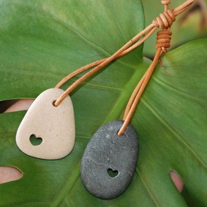 May include: Two stone pendants with heart cutouts on brown leather cords. The stones are a light beige and a dark gray.