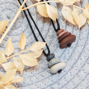 May include: Two necklaces with natural stone pendants rest on a gray, textured surface, alongside dried, light-brown leaves. One pendant features stacked brown stones, the other has a mix of gray and white stones.