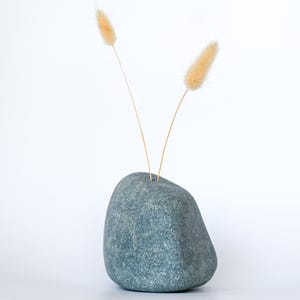 Peut inclure: Un vase en forme de pierre, de couleur bleu-vert, contient deux tiges de fleurs séchées beiges. Le vase a une forme lisse et arrondie, posé sur un fond blanc uni. L'esthétique générale est minimaliste et naturelle.