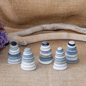 Peut inclure: Cinq sculptures empilées en pierre dans des tons de bleu, gris et blanc, ressemblant à des pyramides miniatures. Chaque sculpture a une pierre sombre au sommet. Le fond présente une surface en toile de jute, une branche sèche et des fleurs violettes.