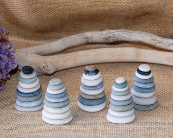 May include: Five stacked stone sculptures in shades of blue, gray, and white, resembling miniature cairns. The sculptures are arranged on a textured, tan surface, with a dried branch and purple flowers in the background.
