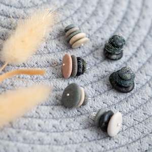 Puede incluir: Primer plano de varios pendientes de piedra con un diseño natural apilado. Los pendientes presentan varios colores, incluyendo gris, beige y rosa, sobre un fondo gris texturizado. Una flor seca añade un toque de elegancia natural.