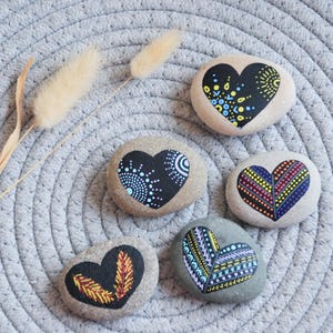 Puede incluir: Cinco piedras en forma de corazón pintadas con diferentes patrones coloridos. Las piedras están dispuestas sobre una superficie circular gris con textura tejida.
