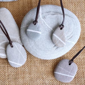 May include: Four gray stone pendants with white veins, each on a brown leather cord. The pendants are displayed on a burlap background.