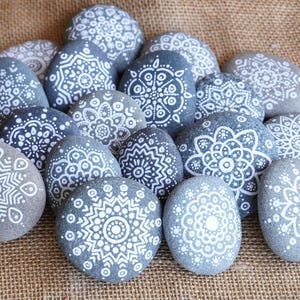 May include: A cluster of grey river stones, each hand-painted with a distinctive white mandala pattern. The stones vary in size and shape, creating a natural and artistic display. The rocks are arranged on a textured, brown surface.