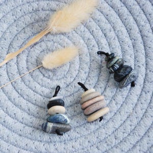 May include: Three decorative stone ornaments with black string loops rest on a light blue woven mat. The ornaments are made of stacked, smooth stones in various colors. A dried, beige plant stem lies across the mat.