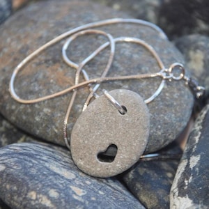 May include: A silver chain necklace with a heart-shaped pendant made from a smooth grey stone. The necklace is displayed on a bed of grey and black stones.