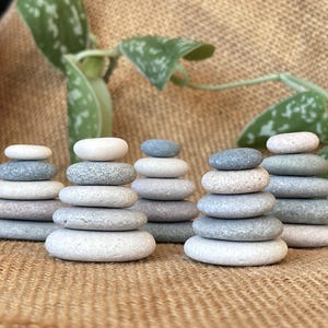 May include: Multiple stacks of smooth, rounded stones in shades of grey and white. Each stack is made up of several stones, creating a balanced, zen-like arrangement. The background features green leaves and a textured, tan surface.