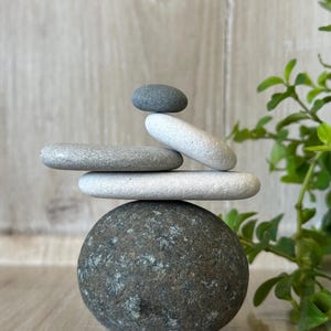May include: A stack of five smooth, rounded stones arranged vertically. The base is a large, dark grey, speckled rock. Above it are two flat, light grey stones, followed by a smaller, tilted light grey stone, and topped with a small, dark grey pebble.