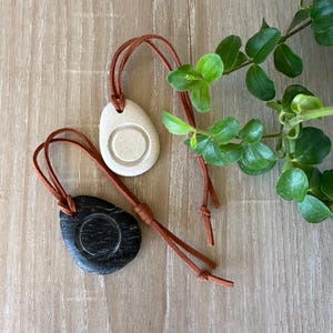 Op de afbeelding: Twee stenen hangers, één crème en één zwart, elk met een cirkelvormige inkeping in het midden. Beide hangers hangen aan dunne, bruine leren koorden die zijn geknoopt om verstelbare lussen te creëren voor gebruik als halsketting.