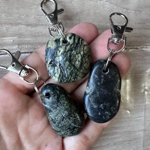 K&ouml;nnte beinhalten: Drei Schl&uuml;sselanh&auml;nger mit glatten, polierten Natursteinen, die an silberfarbenen Karabinerverschl&uuml;ssen aus Metall befestigt sind. Ein Stein ist rund mit gr&uuml;n-schwarzer Maserung, w&auml;hrend die anderen beiden l&auml;nglich sind, einer mit dunkelgr&uuml;ner Marmorierung und einer in einfarbigem Schwarz.
