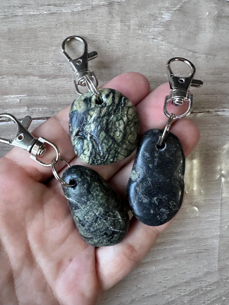 K&ouml;nnte beinhalten: Drei Schl&uuml;sselanh&auml;nger mit glatten, polierten Natursteinen, die an silberfarbenen Karabinerverschl&uuml;ssen aus Metall befestigt sind. Ein Stein ist rund mit gr&uuml;n-schwarzer Maserung, w&auml;hrend die anderen beiden l&auml;nglich sind, einer mit dunkelgr&uuml;ner Marmorierung und einer in einfarbigem Schwarz.