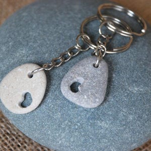 Op de afbeelding: Een set van twee grijze en witte stenen sleutelhangers met hartvormige uitsnijdingen, bevestigd aan een zilveren ketting en sleutelring.