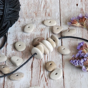 Peut inclure: Un collier fait de perles de pierre naturelle enfilées sur un cordon noir. Les perles sont de différentes nuances de brun et de blanc et ont un trou percé au centre. Le collier est posé sur une surface en bois blanc avec un écheveau de cordon noir et des fleurs séchées.