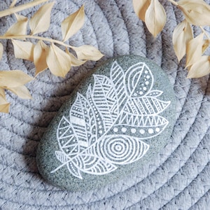 Puede incluir: Una piedra de río lisa, gris y de forma ovalada con un diseño de pluma pintado a mano en color blanco. La pluma está compuesta por intrincados patrones geométricos, incluyendo líneas, puntos y círculos concéntricos en pintura blanca.