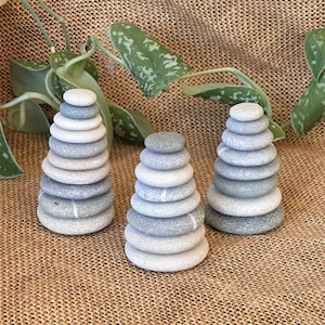 Op de afbeelding: Drie afzonderlijke stapels van gladde, ronde rivierstenen gerangschikt in kegelvormen. Elke stapel bestaat uit meerdere grijze, witte en beige kiezels die op elkaar zijn gebalanceerd en in grootte variëren van de basis tot de top.