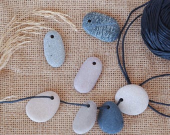 May include: A collection of natural stone pendants in various shapes and colors, including gray, blue, and off-white, with small drilled holes for stringing. A dark blue cord and a bundle of dried grass are also present.