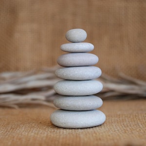 Könnte beinhalten: Ein Stapel glatter, grauer Steine, die übereinander balanciert sind. Die Steine sind in Pyramidenform angeordnet, wobei der kleinste Stein oben liegt. Die Steine liegen auf einer braunen, strukturierten Oberfläche.