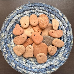 Puede incluir: Una colección de pequeñas piedras planas de color terracota, cada una con un solo agujero perforado, dispuestas en una cesta circular de cuerda trenzada azul y blanca. Las piedras varían en forma y textura, mostrando tonos naranjas y marrones.