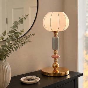 May include: A table lamp with a round white fabric shade, a gold-toned metal base, and a central column featuring a ribbed light blue ceramic cylinder, a small pink ceramic disc, and gold spherical accents, emitting a warm, soft glow.