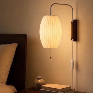 Puede incluir: Una lámpara de pared con una pantalla cilíndrica de tela color blanco roto y textura acanalada. La luz se sostiene mediante un brazo metálico curvo unido a una placa de madera oscura. Una cadena con una pequeña esfera cuelga de la base.