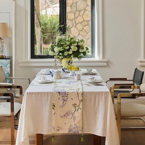 Puede incluir: Una mesa de comedor preparada para una comida, con un mantel blanco y un camino de mesa floral con glicinas moradas. La mesa está puesta con porcelana, una tetera y un jarrón con flores blancas. Sillas doradas rodean la mesa.