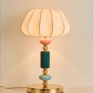 May include: A decorative table lamp with a cream-colored, petal-shaped lampshade. The lamp features a gold base and accents, with a teal, pink, and gold ceramic body. The lamp is designed for indoor use and has a cord.