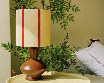 May include: A table lamp with a smooth, dark brown, rounded base and a cylindrical lampshade. The shade is off-white with two vertical, parallel red stripes running down the front. The lamp has a simple, solid structure with a narrow neck.