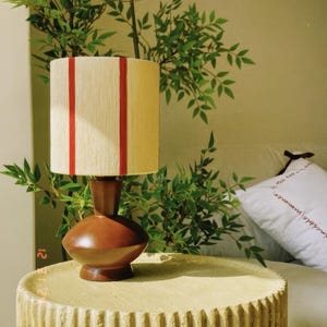 May include: A brown table lamp with a cream-colored shade featuring two vertical red stripes. The lamp sits on a textured, light yellow side table. Green foliage is in the background, creating a natural aesthetic.