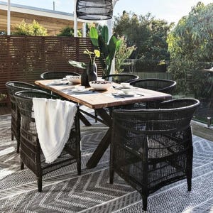 May include: Outdoor dining set featuring a rectangular wooden table and six black wicker chairs. The table is set with white dishes and a centerpiece. A white throw blanket is draped over one chair. The set is on a patterned rug.