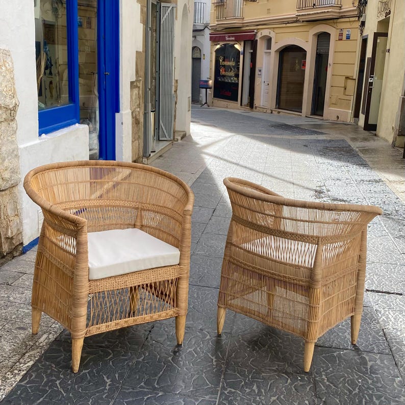 May include: Two wicker armchairs with curved backs and light-colored cushions. The chairs are made of woven natural fibers and have four wooden legs. They are placed on a tiled surface in an outdoor setting.