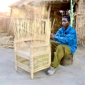 May include: A craftsman weaves a natural fiber chair outdoors. The chair features a high back and armrests, constructed with vertical reeds and woven horizontal supports. The artisan is wearing a blue shirt and khaki pants, seated on a metal stool.