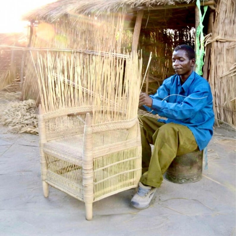 May include: A craftsman weaves a natural fiber chair outdoors. The chair features a high back and armrests, constructed with vertical reeds and woven horizontal supports. The artisan is wearing a blue shirt and khaki pants, seated on a metal stool.