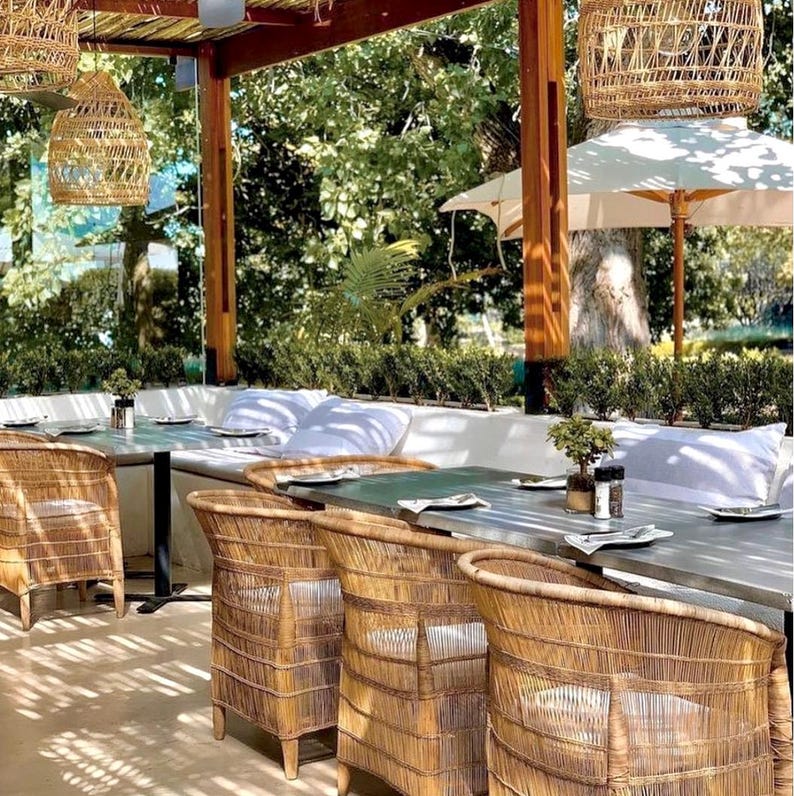 May include: Outdoor dining area with woven rattan chairs and tables. Overhead, woven pendant lights hang from a wooden pergola. White cushions line the built-in bench seating. Green foliage and trees provide a natural backdrop.