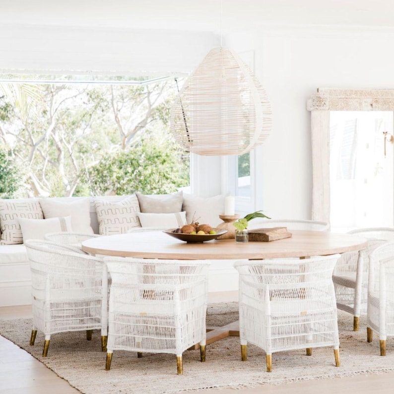 May include: A light wood oval dining table surrounded by white woven chairs with gold-colored legs. A woven pendant light hangs above the table. The room has a neutral color palette with a large window overlooking greenery.