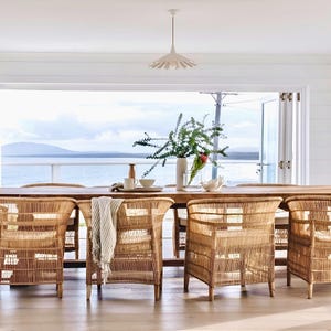 May include: A dining room set with a long wooden table and six woven rattan chairs. A white pendant light hangs above the table, which is set with a vase of greenery, a small bowl, and a decorative piece. The background shows a view of the ocean.