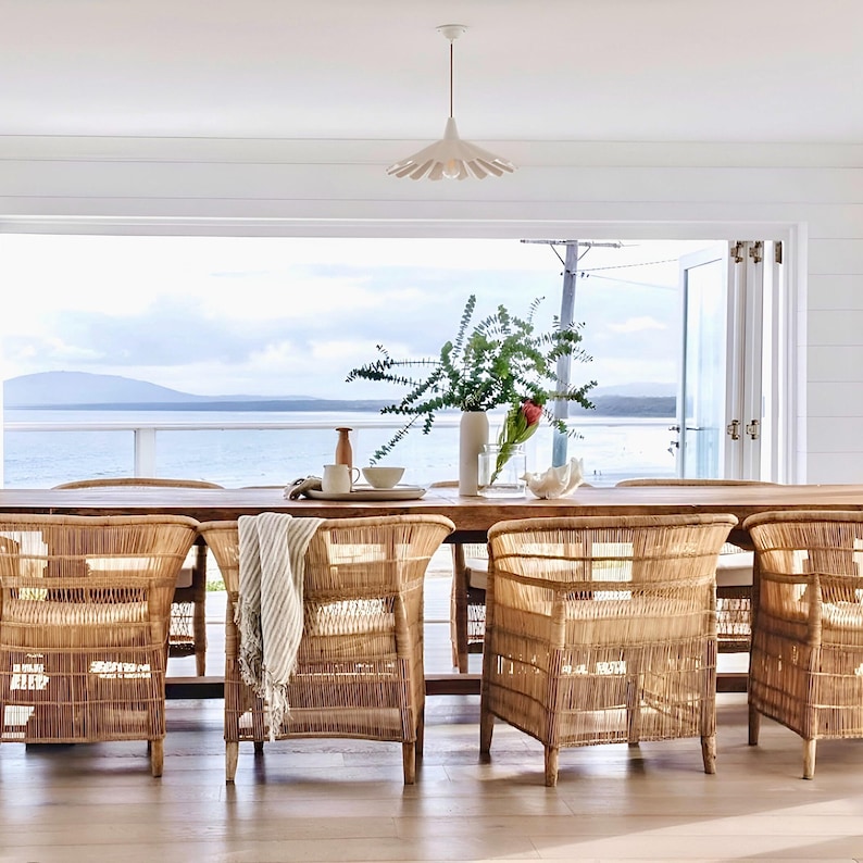 May include: A dining room set with a long wooden table and six woven rattan chairs. A white pendant light hangs above the table, which is set with a vase of greenery, a small bowl, and a decorative piece. The background shows a view of the ocean.