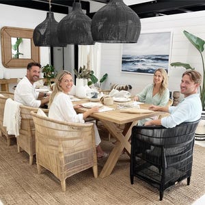May include: A dining room scene featuring a wooden table set for a meal, surrounded by woven chairs. Overhead, three large black woven pendant lights hang. A large painting of the ocean hangs on the wall, and a large rug covers the floor.