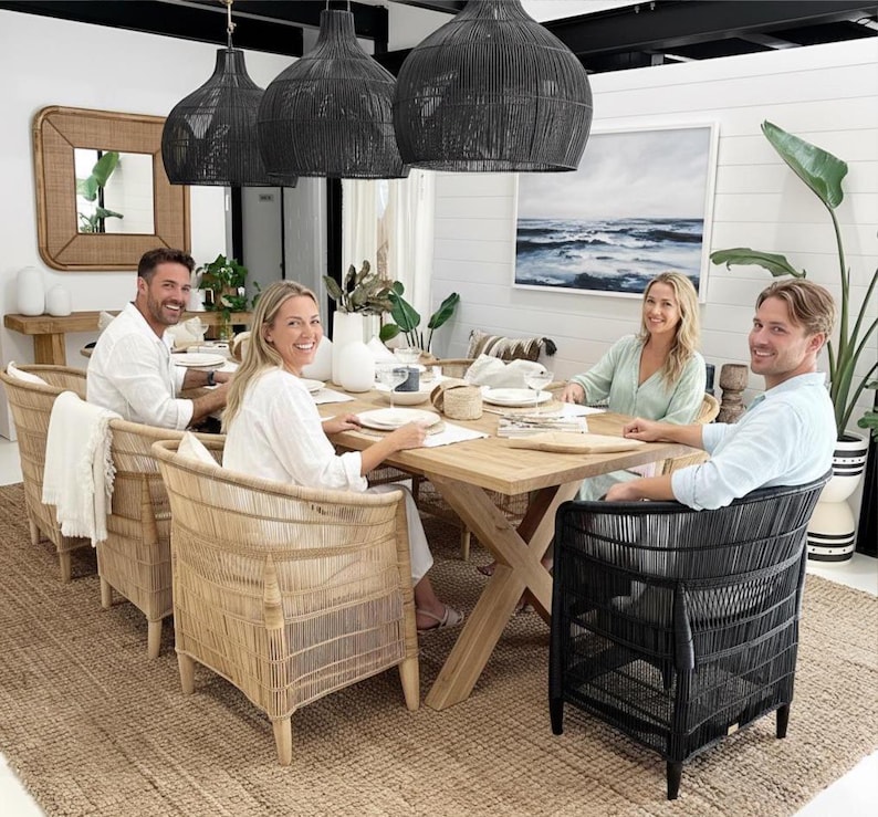 May include: A dining room scene featuring a wooden table set for a meal, surrounded by woven chairs. Overhead, three large black woven pendant lights hang. A large painting of the ocean hangs on the wall, and a large rug covers the floor.