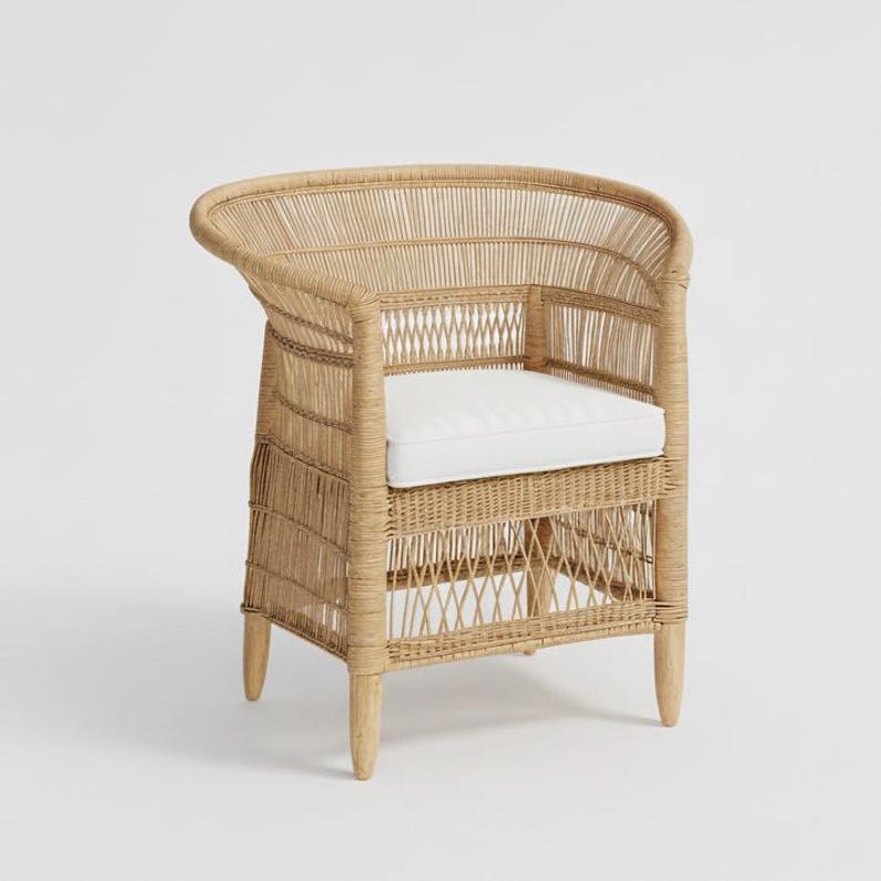 May include: A natural-toned wicker armchair with a curved back and armrests. The chair features a woven design and a white cushion seat. The legs are made of light-colored wood. This chair is suitable for indoor or outdoor use.