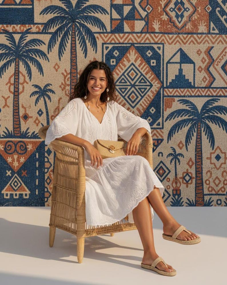 May include: A woman in a white eyelet dress and sandals sits in a wicker chair. She holds a woven clutch. The background features a patterned tapestry with palm trees and geometric designs in blue, tan, and rust.