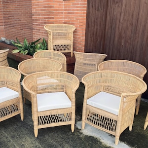 May include: A collection of light brown wicker chairs with white seat cushions. The chairs have curved backs and armrests, and are arranged in a group. The background includes a brick wall and a wooden door.