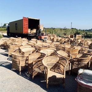 May include: A large outdoor scene featuring numerous woven wicker chairs. The chairs are light brown and have curved backs and armrests. A semi-truck with a shipping container is in the background, with a forklift loading more chairs. The chairs are outdoors on a sunny day.