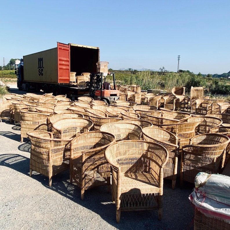 May include: A large outdoor scene featuring numerous woven wicker chairs. The chairs are light brown and have curved backs and armrests. A semi-truck with a shipping container is in the background, with a forklift loading more chairs. The chairs are outdoors on a sunny day.