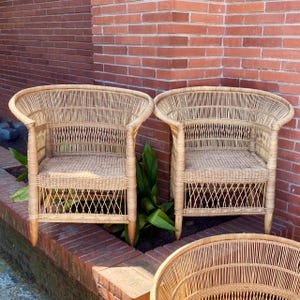 May include: Two natural wicker armchairs with curved backs and woven seats. The chairs have a light brown color and are placed in front of a red brick wall. The chairs have a classic design.
