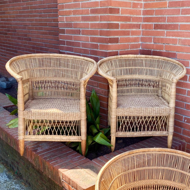 May include: Two natural wicker armchairs with curved backs and woven seats. The chairs have a light brown color and are placed in front of a red brick wall. The chairs have a classic design.