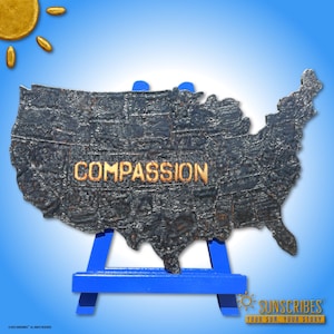 May include: A dark gray metal sculpture of the United States with the word "COMPASSION" in gold lettering. The sculpture is displayed on a small blue easel against a blue gradient background. A golden sun is in the upper left corner.